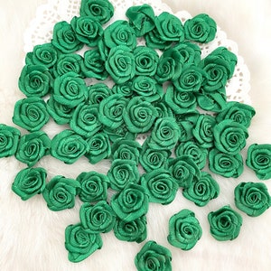 May include: A large quantity of green satin fabric roses. The roses are arranged in a random pattern on a white surface.