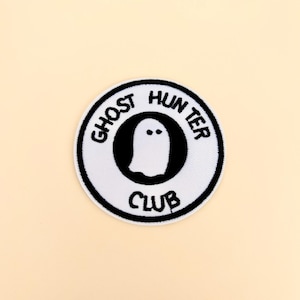 May include: A circular patch with the words "GHOST HUNTER CLUB" in black lettering. The patch features a white ghost illustration inside a black circle, set against a cream-colored background.