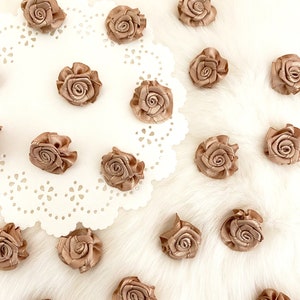 May include: A collection of small, tan-colored fabric roses scattered on a white, lace-edged doily and a fluffy white surface. The roses are tightly wound, with visible petal details. The overall aesthetic is delicate and suitable for craft projects.