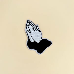 May include: Embroidered patch featuring praying hands. The hands are depicted in white thread with black outlines and fingernails, set against a black base. The patch has a white border and is on a cream-colored background.