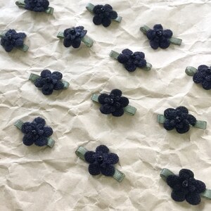 May include: A collection of 12 black fabric flower hair clips with green ribbon accents. The flowers are arranged on a crumpled brown paper background.