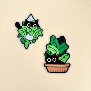 May include: Two embroidered patches featuring black cats amongst green foliage. One cat peeks out from a white pot, the other from a brown planter. The patches have a light blue border and are set against a cream background.