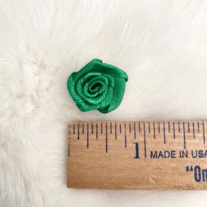 May include: A small green satin rose measuring approximately 1 inch in diameter. The rose is shown on a white fuzzy surface next to a wooden ruler.