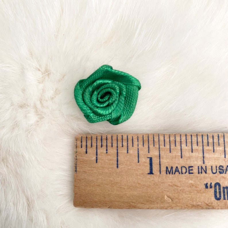 May include: A small green satin rose measuring approximately 1 inch in diameter. The rose is shown on a white fuzzy surface next to a wooden ruler.