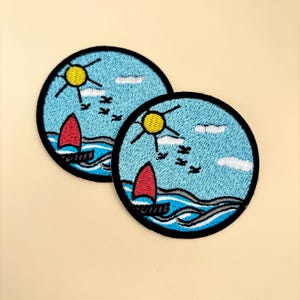 May include: Two embroidered iron-on patches featuring a sailboat with a red sail, a yellow sun, white clouds, and birds flying in a blue sky. The patches are circular and have a black border.