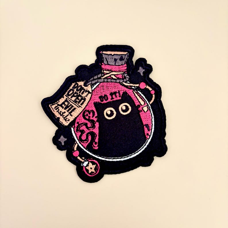 May include: Embroidered patch featuring a black cat inside a potion bottle with the text "DO IT!" and "DON'T OPEN EVIL INSIDE". The patch has a pink and black color scheme with a beige background.