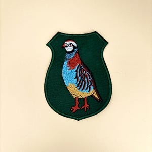May include: Embroidered patch featuring a colorful bird design. The bird has red, blue, and brown feathers, with a yellow beak and legs. The patch is shield-shaped with a dark green border, suitable for jackets, bags, or other fabric items.