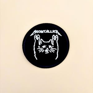 May include: A round, black patch with the white text "MEOWTALLICA" above a cat silhouette with raised paws. The cat's face has whiskers and a small nose. The patch is likely for clothing or accessories.
