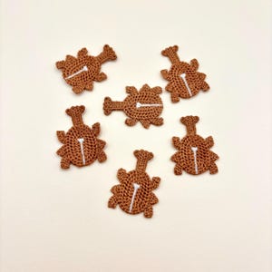May include: Six brown crocheted appliques shaped like chicken drumsticks. Each piece features a textured, yarn-based surface with a single vertical white embroidered line running through the center, mimicking the appearance of a bone.