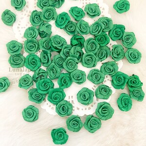 May include: A collection of small, emerald green satin ribbon roses scattered on a white, lacy surface. The roses are tightly wound, with visible petal details. The image is well-lit, highlighting the texture and color of the floral embellishments.