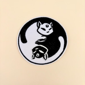 May include: A circular embroidered patch featuring a black and white yin-yang design composed of two stylized cats. The white cat is positioned on the black side, and the black cat is on the white side, both with closed eyes and a crescent moon symbol.