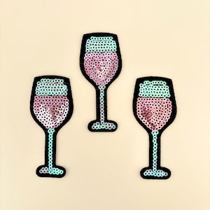 May include: Three sequined wine glass patches. Each patch features a black outline, a pink sequined bowl, and a turquoise sequined rim. The stems and bases are also black.