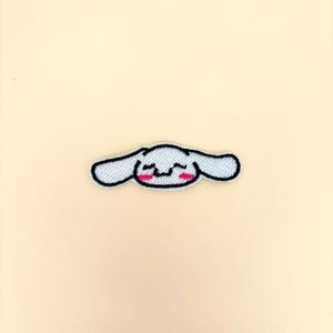 Peut inclure: Écusson brodé représentant un personnage de dessin animé avec un corps blanc, de longues oreilles et un contour noir. Le personnage a un visage souriant avec des joues roses. L'écusson est sur un fond beige clair.