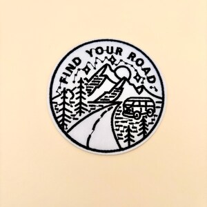 May include: A circular patch with a black and white design. The patch features a road winding through a mountain landscape with trees, a sun, and a van. The text "FIND YOUR ROAD" is arched around the top.