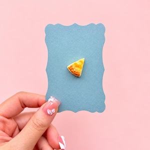 May include: A small, triangular-shaped charm resembling a slice of golden-brown pie, centered on a scalloped-edge, slate-blue rectangular card. The pie slice features a textured, baked crust appearance with a warm yellow and brown color palette.