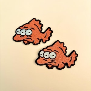 Op de afbeelding: Twee oranje geborduurde vispatches met drie lichtblauwe ogen en zwarte contouren. De vissen hebben een cartoonachtig ontwerp en staan tegen een cr&egrave;mekleurige achtergrond. Geschikt voor kleding of accessoires.