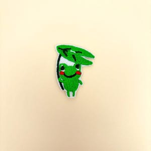 May include: Embroidered patch featuring a smiling green frog holding a green leaf. The frog has red cheeks and a white outline. The patch is on a cream-colored background. This patch is suitable for clothing or accessories.