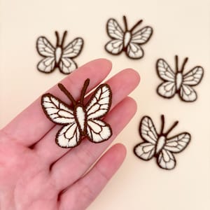 May include: Embroidered butterfly patches in brown and white. The patches feature detailed wing patterns and are arranged on a light background. These patches are ideal for adding a decorative touch to clothing, bags, or other fabric items.