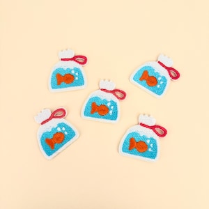 Peut inclure: Cinq écussons brodés en forme de petit sac en plastique rempli d'eau bleue avec un poisson rouge. Les écussons présentent des sommets blancs avec des liens rouges, des sections d'eau bleu clair et des poissons orange avec des bulles blanches.