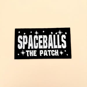 May include: Black rectangular patch with white embroidered text that reads "SPACEBALLS" above "THE PATCH" with star accents. The patch is likely for a jacket, bag, or other apparel item.