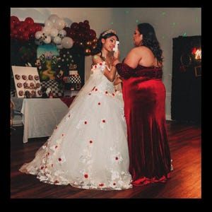 May include: Two people, one wearing a white gown with red flowers and a crown, the other wearing a red velvet dress, are embracing. The person in the white gown is crying. The background is decorated with red and white balloons.