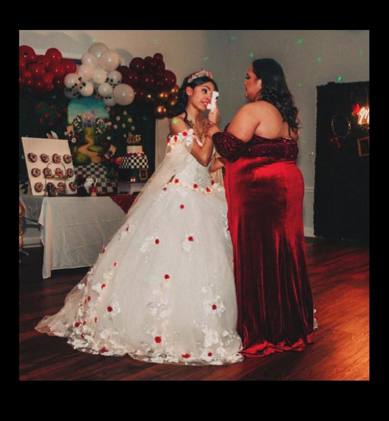 May include: Two people, one wearing a white gown with red flowers and a crown, the other wearing a red velvet dress, are embracing. The person in the white gown is crying. The background is decorated with red and white balloons.