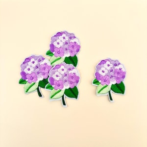 May include: Four purple and white embroidered flower patches with green leaves. The patches are on a light beige background.