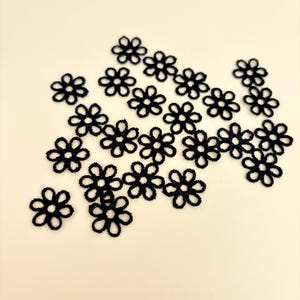 May include: A collection of small, black, openwork flower-shaped embellishments. These delicate floral accents are scattered across a light-colored surface. The intricate details of each flower are clearly visible, showcasing a craft or sewing supply.