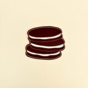 May include: A dark brown embroidered patch in the shape of a stack of cookies with white filling. The patch has a stitched border and is set against a cream-colored background. The cookies appear to be a popular snack.
