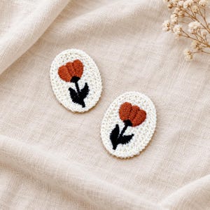 May include: Two oval-shaped patches featuring white crocheted borders and centers. Each patch displays an embroidered design of a rust-colored flower head with a black stem and leaves centered on the white fabric background.