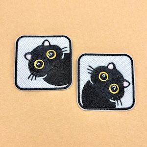 May include: Two square embroidered patches featuring a black cat with large, wide yellow eyes against a light blue background. Each patch has a black border and depicts the cat looking upward with a curious expression and visible white whiskers.