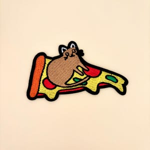 May include: Embroidered patch featuring a brown cat lounging on a slice of pizza. The pizza has a yellow crust, red sauce, and green and red toppings. The patch has a black border.