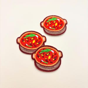 May include: Three circular embroidered patches featuring a red bowl filled with a stew containing yellow and dark red ingredients, topped with green scallions. Each patch has a brown base and a dark brown border with two small side handles.