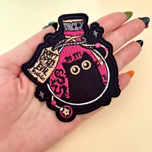May include: Embroidered patch featuring a black cat inside a pink potion bottle. The patch includes the text "DON'T OPEN EVIL inside" and "DO IT!" in white lettering. The patch is outlined in black and white.