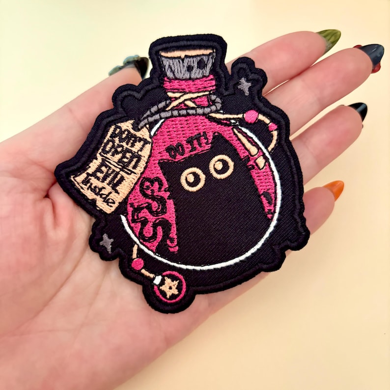 May include: Embroidered patch featuring a black cat inside a pink potion bottle. The patch includes the text "DON'T OPEN EVIL inside" and "DO IT!" in white lettering. The patch is outlined in black and white.