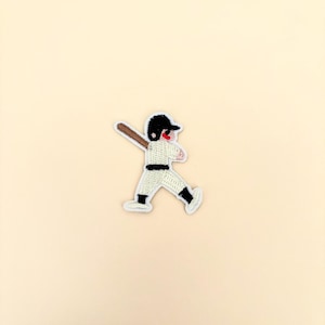 May include: Embroidered patch featuring a baseball player in a batting stance. The patch has a white uniform with black accents, a black helmet, and a brown bat. The player's face is red. The patch is on a cream-colored background.