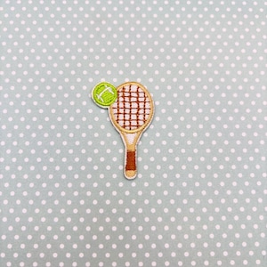May include: Embroidered patch featuring a tennis racket and ball. The racket is tan with a brown handle and brown cross-strings. The tennis ball is green with white stitching. The patch is on a light blue background with white polka dots.