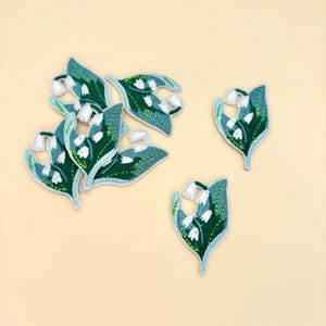 May include: Embroidered patches featuring lily of the valley flowers. The patches have a teal and green leaf design with white flower details. The patches are arranged on a light beige background.