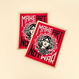 May include: Two red rectangular patches with white text and a black and white illustration of a woman's face. The text reads "MAKE ART" at the top and "NOT WAR" at the bottom.