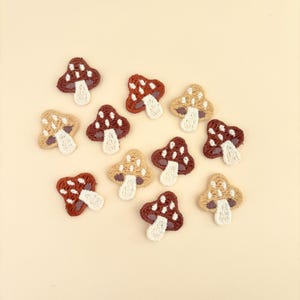 May include: A collection of embroidered mushroom patches in various shades of brown, tan, and white. The mushroom caps are dotted with white, and the stems are white. The patches are arranged on a light beige background.