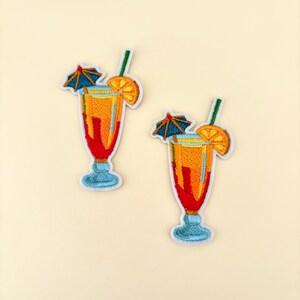 May include: Two embroidered patches shaped like tropical cocktails. Each patch features a tall glass with orange, red, and yellow colors, a blue base, a green straw, a slice of orange, and a small blue umbrella. The patches are on a light beige background.