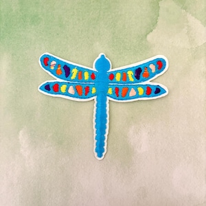 May include: A blue embroidered dragonfly with colorful accents. The dragonfly has a long, thin body and two pairs of wings.
