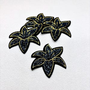 May include: Four black flower patches with gold glitter accents. Each patch has a detailed floral design with a black base and gold glitter outlining the petals. The patches are arranged on a white textured surface.