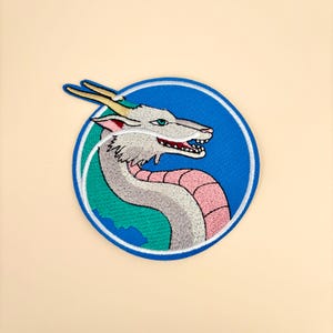 May include: Embroidered patch featuring a white dragon with gold horns and a pink and white body. The dragon is set against a blue circle with a white border, and a green background. The patch is on a beige background.