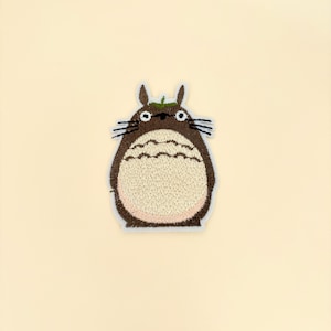 May include: Embroidered patch featuring a cartoon character with a brown body, tan belly, and black whiskers. The character has small ears and a green leaf on its head. The patch has a white border and is set against a cream-colored background.