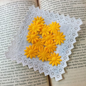 May include: A close-up of a white lace doily with yellow felt flower embellishments. The doily is on top of a book with text.