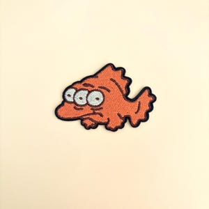 Op de afbeelding: Geborduurde oranje vispatch met drie ogen en een chagrijnige uitdrukking. De patch heeft een zwarte rand en is geschikt voor kleding of accessoires. Het visontwerp is in cartoonstijl.