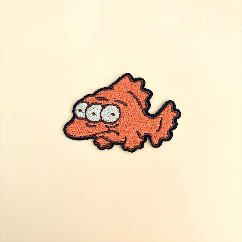 Op de afbeelding: Geborduurde oranje vispatch met drie ogen en een chagrijnige uitdrukking. De patch heeft een zwarte rand en is geschikt voor kleding of accessoires. Het visontwerp is in cartoonstijl.