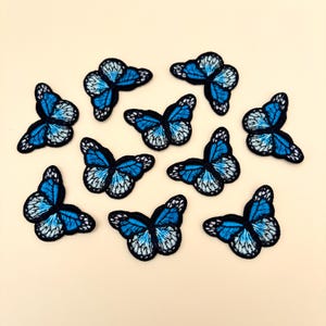 May include: Ten blue and white butterfly iron-on patches. The butterflies have black outlines and are arranged in a scattered pattern on a light beige background.