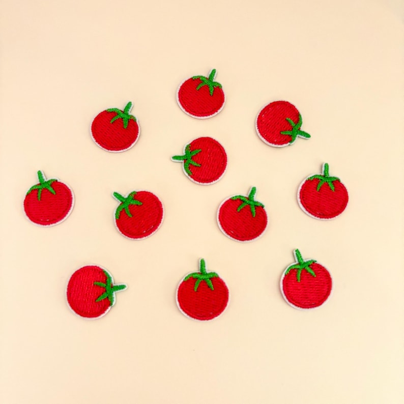 Pode incluir: Uma cole&ccedil;&atilde;o de onze patches de tomate bordados. Cada patch apresenta um tomate vermelho vibrante com um caule verde e uma borda branca. Os patches est&atilde;o dispostos sobre um fundo bege claro.